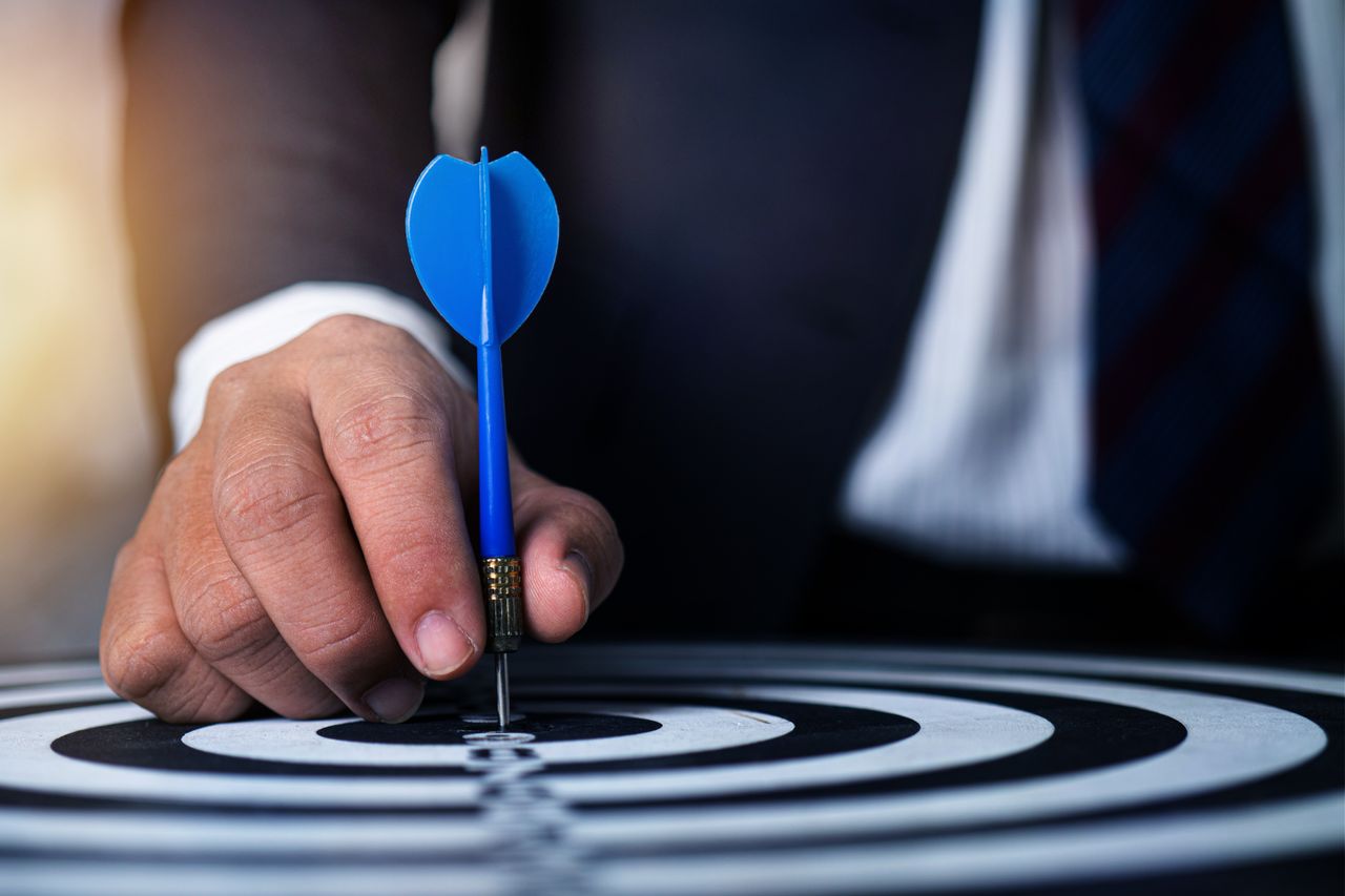 Services Close-up of a man's hand holding a blue dart on a bullseye, symbolizing precision and focus.